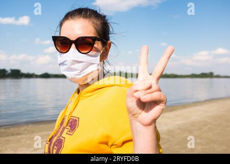 Jeune femme portant un masque facial debout près de la rivière dans la nature montrant le geste de doigt de victoire de V Banque D'Images