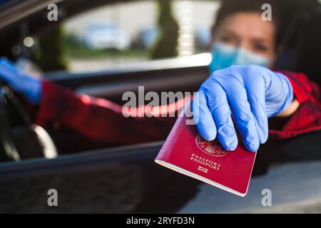 Jeune femme conductrice portant un masque de protection et des gants donnant passeport rouge par la fenêtre de la voiture Banque D'Images