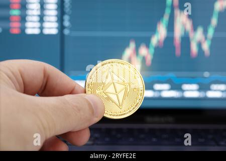 Main tenant une pièce de monnaie Ethereum et un tableau sur l'écran de l'ordinateur portable en arrière-plan.Concept de négociation ou d'investissement de crypto-monnaie Banque D'Images