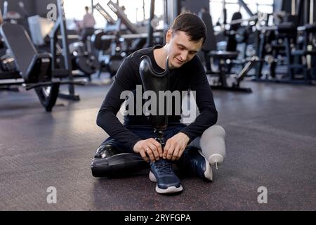 souriant jeune beau sportif avec la jambe artificielle assis sur le sol à la salle de gym. style de vie, temps libre, temps libre, motivation Banque D'Images