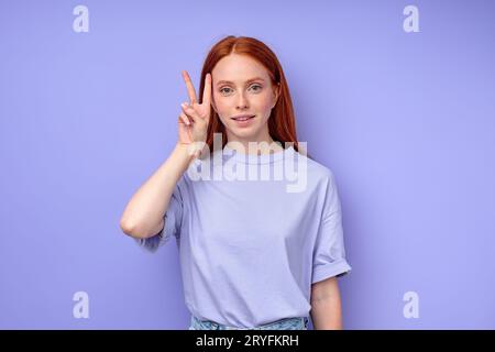 jolie fille rousse montrant deux doigts regardant la caméra, signe de victoire. gros plan portrait isolé fond bleu. femme démontrant la lette Banque D'Images