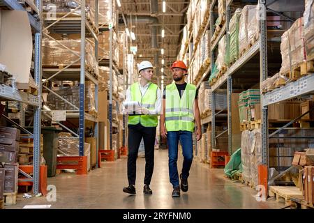 deux hommes ingénieurs en casques de sécurité et uniformes verts marchant parmi les étagères avec des marchandises dans l'entrepôt parlant, vérifiant les marchandises. logistique et affaires ex Banque D'Images