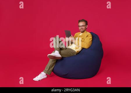 Beau corps pleine grandeur jeune homme 20s, 30s portant des vêtements denim décontractés assis dans un fauteuil sac tenant la tablette numérique dans les mains de travail Banque D'Images