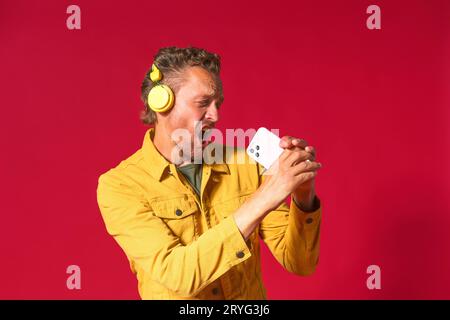 Chantant bel homme appréciant sa chanson préférée à l'aide du téléphone et des écouteurs sans fil portant un Jean veste jaune isolé sur le rouge Banque D'Images