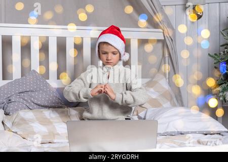 En attente d'un miracle de Noël. Un jeune enfant d'âge préscolaire dans un chapeau de santa communique via un lien vidéo expliquant le cadeau qu'il veut recevoir dans un décora de chambre Banque D'Images