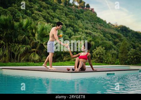 Couple profitant de l'après-midi d'été au bord de la piscine : homme caucasien en bermuda offrant du jus d'orange à sa copine afro-américaine dans une épaule Banque D'Images