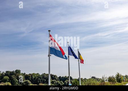 Zone frontalière avec trois drapeaux sur des mâts de drapeau avec des arbres verts feuillus et ciel bleu brumeux en arrière-plan, drapeaux de la Hollande, de l'Union européenne et de la Belgique, su Banque D'Images