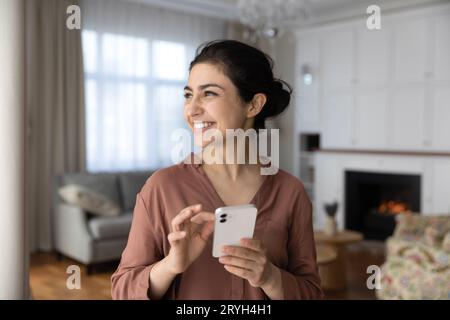 Femme indienne souriante rêveuse tenant smartphone, regardant dans la distance Banque D'Images