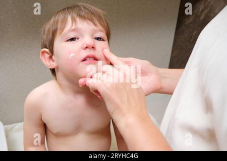 La mère de femme frotte ses joues d'enfant avec de la crème d'allergie dans la salle de bain de la maison. Enfant âgé de deux ans (garçon de deux ans) maman met de la crème sur le visage de bébé garçon wi Banque D'Images