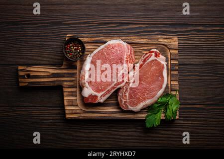 Couper les steaks de porc crus avec des assaisonnements sur la planche à découper de cuisine, fond en bois rustique d'en haut, prêt pour le barbecue. Longe de porc Banque D'Images