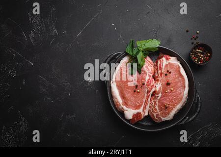 Couper les steaks de porc crus avec des assaisonnements dans une poêle en fonte noire, fond de pierre rustique sombre vue de dessus, prêt à rôtir. Porc Banque D'Images