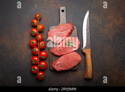 Trois steaks d'aloyau de bœuf crus non cuits sur une planche à découper en bois avec romarin sur une planche à découper en bois avec tomate cerise Banque D'Images