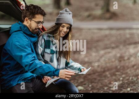 Positive jeune famille caucasienne dans des vestes assis dans le coffre de voiture, regardez la carte de route, profitez de l'aventure le week-end Banque D'Images