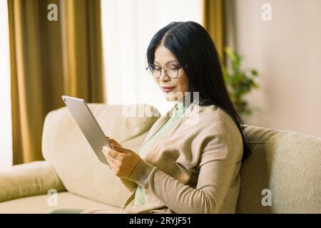 Femme asiatique mature portant des lunettes est profondément axée sur la navigation sur Internet sur sa tablette tout en étant confortablement assis dans l'inte Banque D'Images