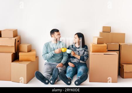 Jeune couple prenant une pause le jour de déménagement assis sur le sol et célébrant leur nouvelle vie avec boisson Banque D'Images