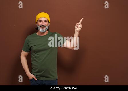 Homme européen souriant dans un chapeau jaune pointe sa main sur le côté avec un espace de copie sur un fond brun. Placement de produit, adverti Banque D'Images