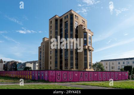 L'écriture et les images peintes de couleurs vives déclarant que vous êtes aimé sur un mur entourant et un immeuble d'appartements à Portsmouth. Sept. 2023 Banque D'Images