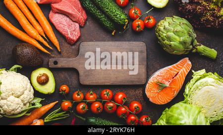 Divers produits alimentaires crus : légumes, viande de boeuf, saumon de poisson et planche à découper vide en bois au centre, pierre brun rustique foncé Banque D'Images
