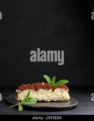 Un morceau de tiramisu arrosé de cacao, sur une branche de menthe fraîche sur un fond noir Banque D'Images