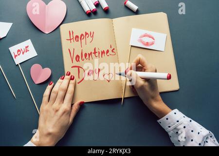 Attributs de la Saint-Valentin sur un bureau gris. La femme écrit un souhait pour la Saint-Valentin dans un livre de croquis. Banque D'Images