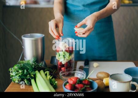 Une jeune femme en tablier à la cuisine met les ingrédients pour un smoothie dans un bol mélangeur. Concept de saine alimentation. Banque D'Images