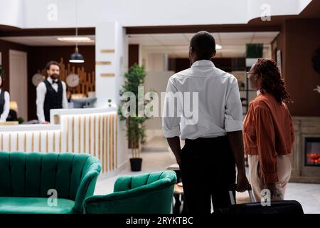 Les personnes arrivant dans le hall de l'hôtel de luxe, entrant dans le salon et se préparant à faire le processus d'enregistrement. Jeune couple parlant aux employés de la réception pour la réservation de chambre, comptoir de réception. Banque D'Images