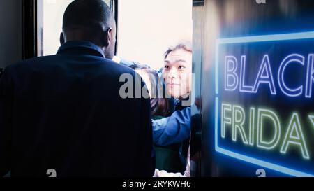 Les gens qui deviennent fous pour entrer à l'intérieur du magasin d'abord pour trouver des vêtements à moitié prix, les meilleures offres pendant le Black friday. Foule agressive de clients poussant un agent de sécurité, attendant à l'entrée du centre commercial. Banque D'Images