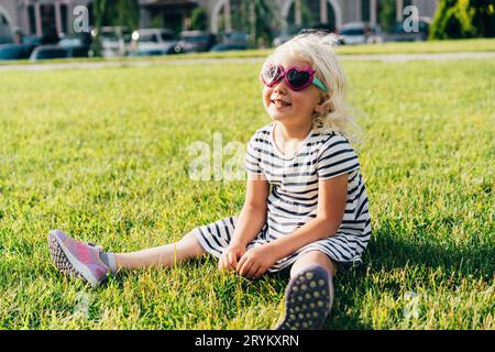 Une petite fille souriante et heureuse s'assoit sur une pelouse dans un environnement urbain. Un enfant content dans un été en appréciant l'humeur. Banque D'Images