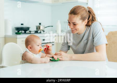 Heureuse maman rousse souriante nourrit son bébé avec de la purée de légumes dans la cuisine à la maison. Routine quotidienne de garde et d'éducation des enfants Banque D'Images