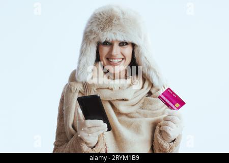 heureuse femme moderne d'âge moyen en manteau d'hiver et chapeau de fourrure sur fond blanc dans des gants blancs avec carte de crédit et smartphone. Banque D'Images