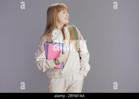 fille moderne souriante en survêtement beige avec cahiers et écouteurs contre gris. Banque D'Images