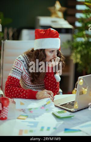 Noël. Femme de propriétaire de petite entreprise moderne stressée dans le chapeau de santa et pull de Noël rouge avec des documents et un ordinateur portable travaillant dans le vert moderne de Banque D'Images