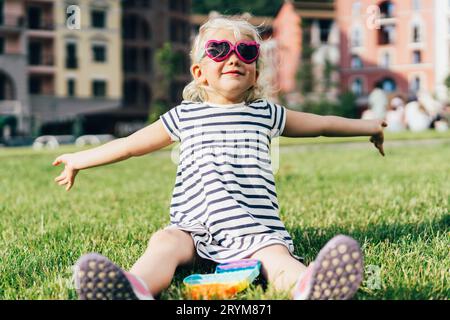 Une petite blonde drôle de lunettes de soleil et une robe rayée se trouve sur la pelouse. Banque D'Images