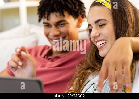 Joyeux couple divers utilisant une tablette se détendre sur le canapé dans le salon et rire Banque D'Images