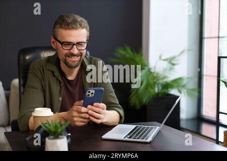 Freelance positive qui travaille dans le confort de la maison. Homme souriant a vu un message texte au téléphone tout en prenant une pause bien méritée Banque D'Images