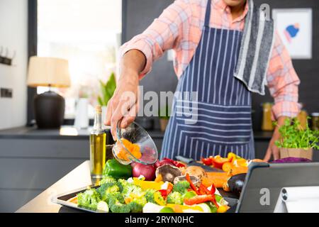Section centrale de l'homme biracial portant un tablier de cuisine repas, verser des légumes dans la cuisine Banque D'Images