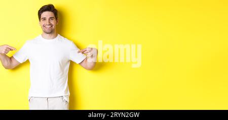 Joyeux gars attrayant pointant des doigts sur votre logo, montrant la promotion sur son t-shirt, debout sur fond jaune Banque D'Images