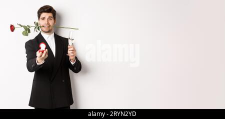 Romantique jeune homme en costume faire une proposition, tenant rose dans les dents et un verre de champagne, montrant l'anneau d'engagement, demandant à M. Banque D'Images