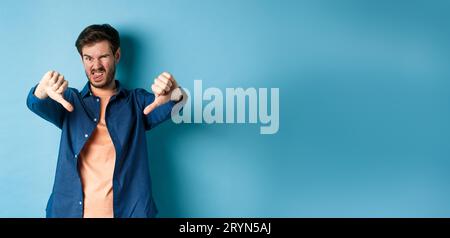 Un gars dégoûté exprime une opinion négative, montrant les pouces vers le bas et la langue, fronçant les sourcils bouleversés, désapprouvant quelque chose de mauvais, debout sur bl Banque D'Images