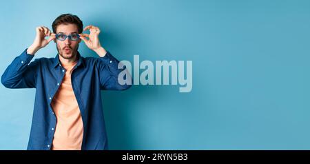 Drôle jeune homme essayant de nouvelles lunettes de soleil, regardant surpris à l'appareil photo, debout sur fond bleu Banque D'Images