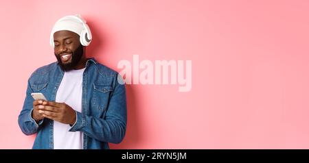 Bel homme noir élégant écoutant de la musique dans des écouteurs et écrivant un message sur smartphone, souriant heureux, debout sur rose ba Banque D'Images