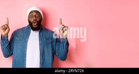 Enthousiaste et impressionné Black man montrant une incroyable promo, pointant les doigts vers le haut du logo, debout sur fond rose Banque D'Images