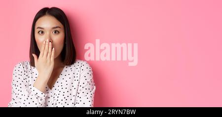 Image d'une jeune fille asiatique choquée commérant, étouffant et couvrant la bouche, regardant la caméra avec incrédulité complète, debout sur le dos rose Banque D'Images