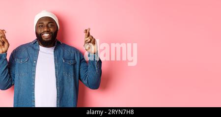 Homme afro-américain optimiste faisant des voeux, tenant les doigts croisés et souriant, debout sur fond rose Banque D'Images