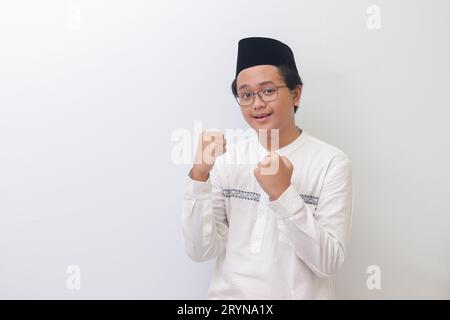 Portrait d'un jeune homme musulman asiatique levant le poing, célébrant le succès et regardant la caméra. Image isolée sur fond blanc Banque D'Images