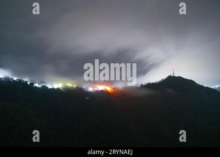 Prise de vue nocturne longue montrant le brouillard et les nuages roulant sur les montagnes avec des lumières bleues et oranges du village de Darjeeling, Shimla, manali Banque D'Images