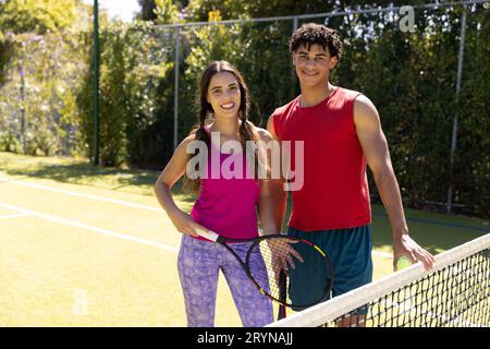 Portrait de jeune couple biracial souriant avec raquettes de tennis debout par filet sur le terrain contre les arbres Banque D'Images