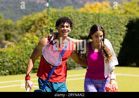 Jeune couple biracial joyeux avec les armes autour de marche hors du court de tennis après le match Banque D'Images