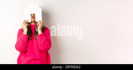 Immobilier. Drôle asiatique femme d'âge moyen cachant le visage derrière le modèle de maison de papier et montrant des lèvres plissées, s'amusant, debout Banque D'Images