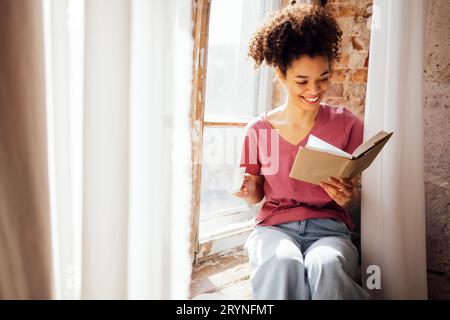 Jeune souriant jolie fille de race mixte est assise sur le rebord de la fenêtre avec tasse de thé en porcelaine et livre. Banque D'Images